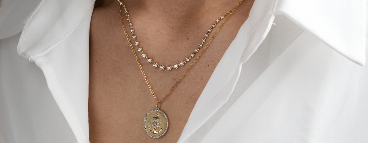 A close-up of a person wearing layered necklaces over a white collared shirt. The necklaces include a gold chain with a round pendant and a beaded pearl necklace.