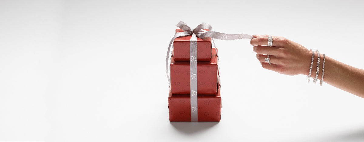 Three stacked red gift boxes tied with a silver ribbon and bow, with a person's hand reaching in from the right against a clean white background.